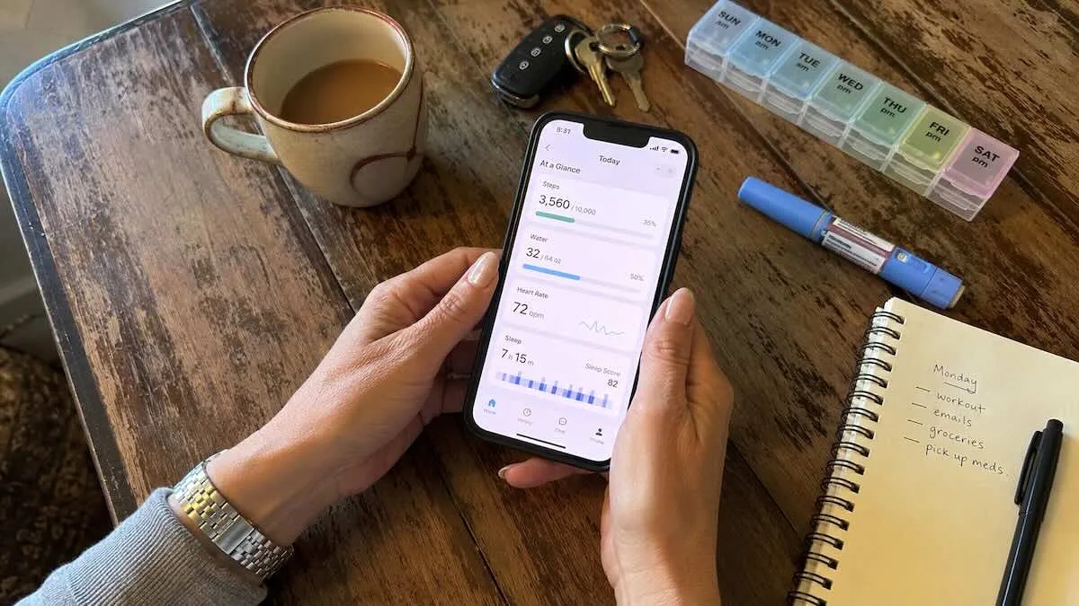 Hands holding a phone with a health-tracking app open, beside a GLP-1 injection pen and a weekly pill organizer.